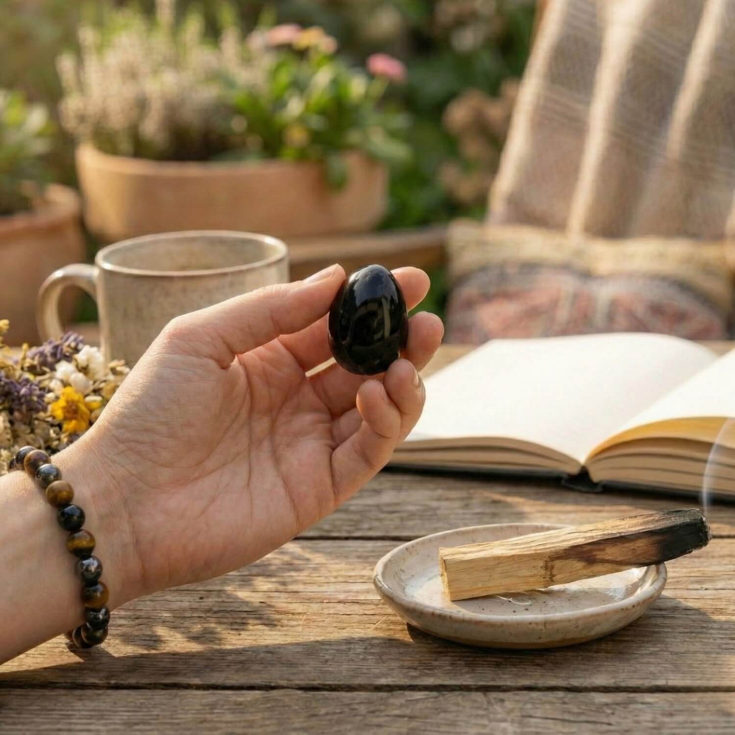 Mano sosteniendo un huevo de Obsidiana Negra durante un ritual de limpieza con humo de Palo Santo. La muñeca luce la pulsera de Triple Protección (Ojo de Tigre, Hematita, Obsidiana).