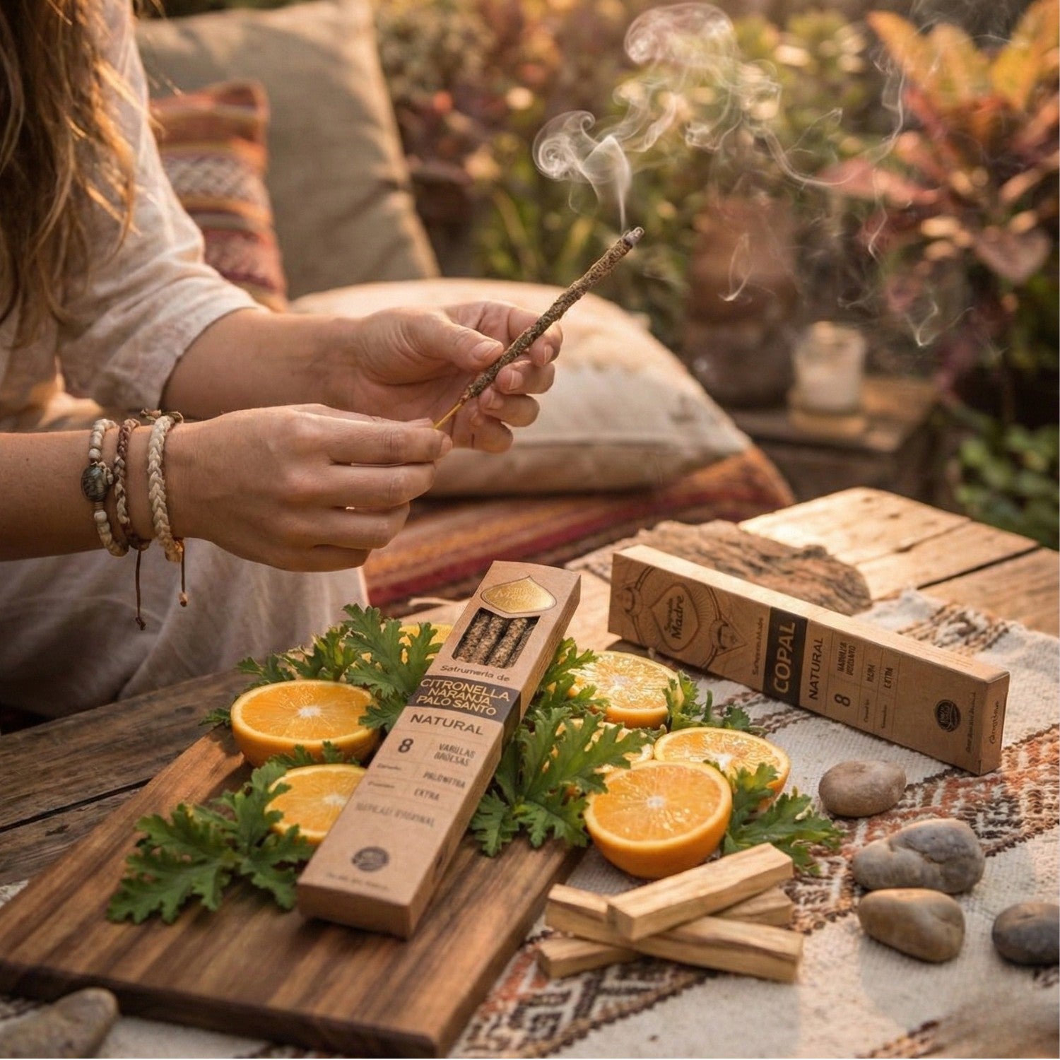 Ritual al aire libre con Pack de Inciensos Naturales Sagrada Madre. Manos encendiendo una varilla junto a cajas de Copal y Citronella con Naranja y Palo Santo sobre mesa de madera.