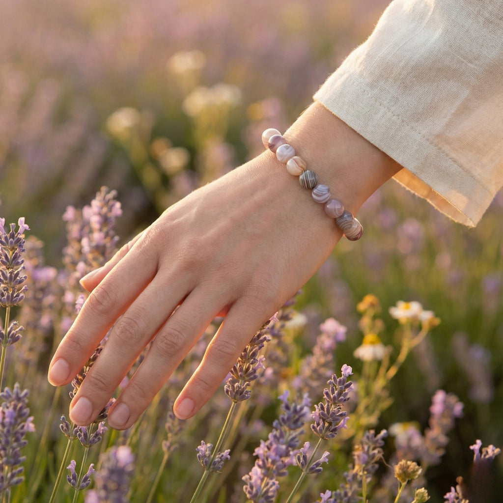 Mano de mujer acariciando flores de lavanda luciendo una pulsera de Ágata de Botsuana. Joyería natural en tonos grises y rosados para la paz interior y el consuelo.