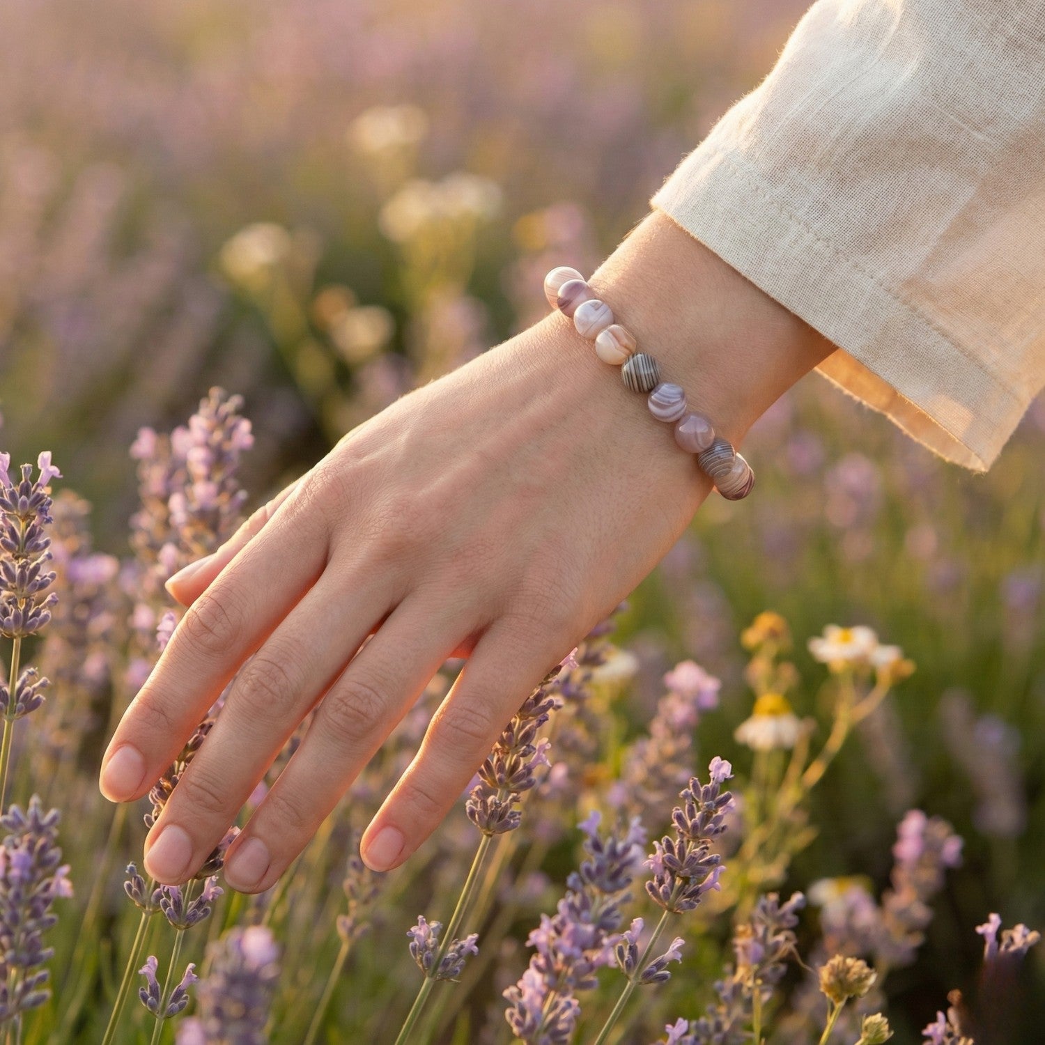Mano de mujer acariciando flores de lavanda luciendo una pulsera de Ágata de Botsuana. Joyería natural en tonos grises y rosados para la paz interior y el consuelo.